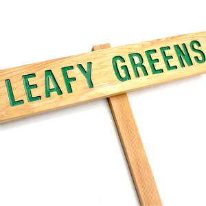 Op de afbeelding: Een houten bord met de woorden "LEAFY GREENS" in groene letters. Het bord is bevestigd aan een houten paal.