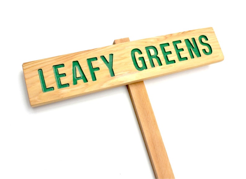 Op de afbeelding: Een houten bord met de woorden "LEAFY GREENS" in groene letters. Het bord is bevestigd aan een houten paal.