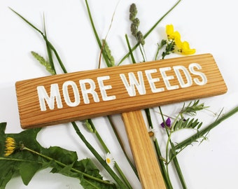 Puede incluir: Un letrero de madera con las palabras "MORE WEEDS" pintadas en blanco. El letrero está rodeado de varios tipos de malas hierbas verdes y flores silvestres, incluidas flores amarillas y flores moradas.