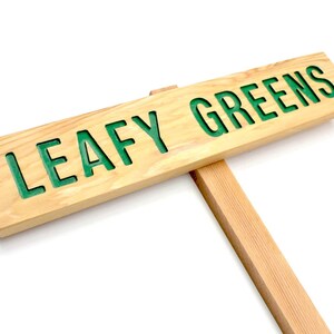 Op de afbeelding: Een houten bord met de tekst "LEAFY GREENS" in groene letters. Het bord is bevestigd aan een houten paal.