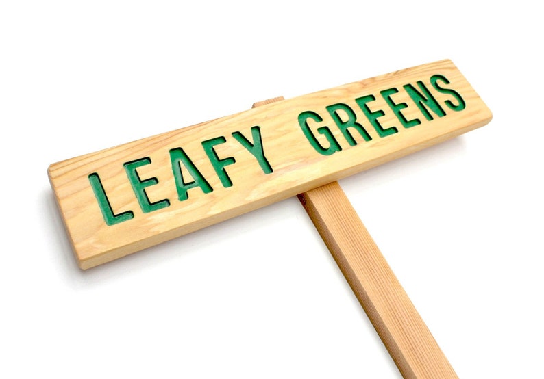 Op de afbeelding: Een houten bord met de tekst "LEAFY GREENS" in groene letters. Het bord is bevestigd aan een houten paal.