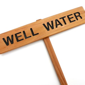 May include: A wooden sign with the text "WELL WATER" in black lettering. The sign is attached to a wooden stake.