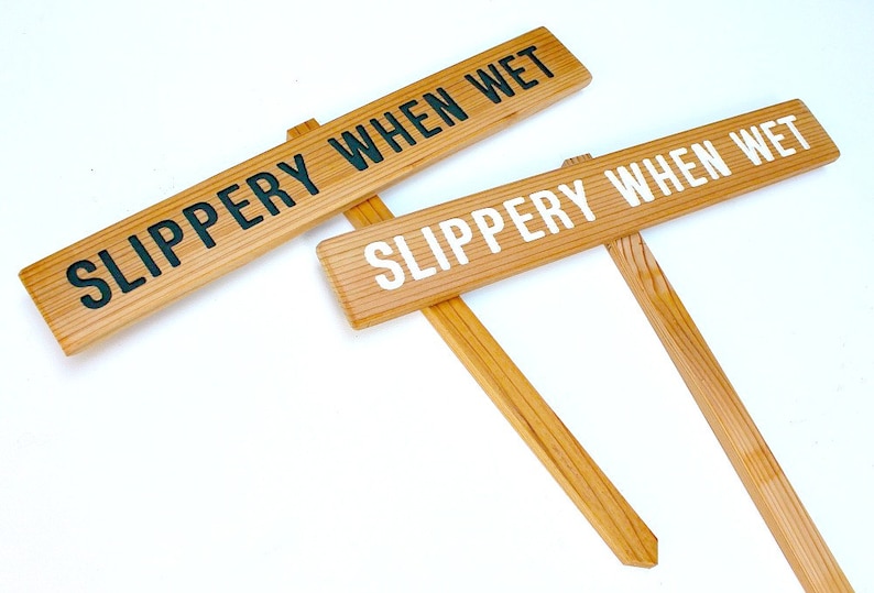 May include: Two wooden signs with the text "SLIPPERY WHEN WET" in black and white lettering. The signs are attached to wooden stakes.