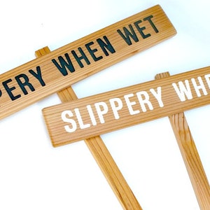 May include: Two wooden signs with the text "SLIPPERY WHEN WET" in black and white lettering. The signs are attached to wooden stakes.