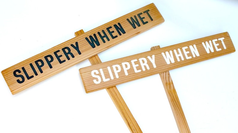 May include: Two wooden signs with the text "SLIPPERY WHEN WET" in black and white lettering. The signs are attached to wooden stakes.