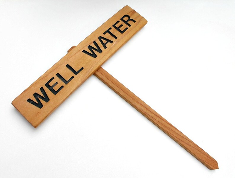May include: A wooden sign with the text "WELL WATER" in black lettering. The sign is attached to a wooden stake.