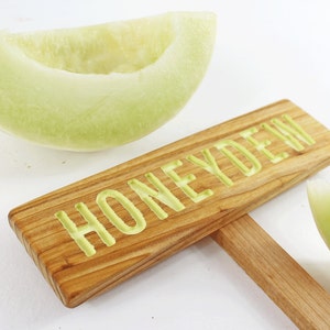 Może przedstawiać: Drewniany znak z wygrawerowanym słowem "Honeydew" w jasnozielonym kolorze. Znak jest podparty drewnianym patykiem. Obok znaku znajduje się plaster melona miodowego.