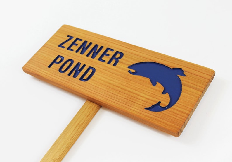 Może przedstawiać: Drewniany znak z napisem "Zenner Pond" w niebieskich literach. W znak wyrzeźbiono niebieską sylwetkę ryby.
