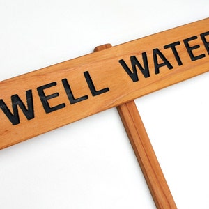 May include: A wooden sign with the text "WELL WATER" in black lettering. The sign is attached to a wooden stick.
