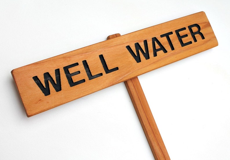 May include: A wooden sign with the text "WELL WATER" in black lettering. The sign is attached to a wooden stick.