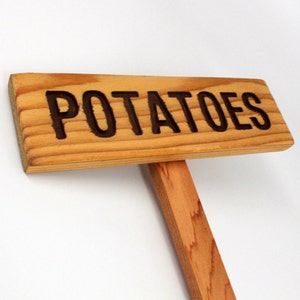 May include: A wooden sign with the word "POTATOES" carved into it. The sign is attached to a wooden stake.