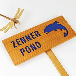 Może przedstawiać: Drewniany znak z napisem "Zenner Pond" w niebieskich literach. W znaku wyrzeźbiona jest niebieska sylwetka ryby.