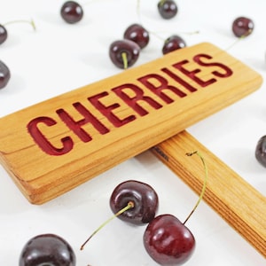Może przedstawiać: Drewniany znak z wygrawerowanym słowem "CHERRIES" czerwonymi literami. Znak jest podparty drewnianym patykiem. Wokół znaku rozrzucone są ciemnoczerwone wiśnie.