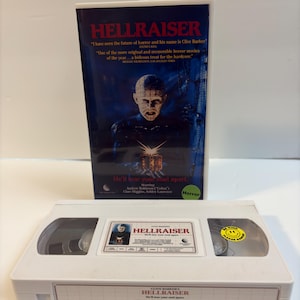 May include: A white VHS tape of the movie Hellraiser with a central label featuring the title and a small image. The tape includes a yellow circular sticker that reads Be Kind Please Rewind. It is displayed in front of its original blue movie box.