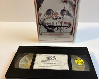 Może przedstawiać: Czarna kaseta VHS i jej białe kartonowe etui do Scary Stories to Tell in the Dark. Etui zawiera ilustrację popękanej, bladej, potwornej twarzy oraz czerwoną naklejkę Horror. Kaseta posiada żółtą naklejkę Be Kind Rewind.