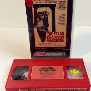 May include: A red VHS cassette tape of The Texas Chain Saw Massacre with a yellow smiley face sticker that reads Be Kind Rewind, positioned in front of its original clear plastic case featuring a poster with red and black text and imagery.