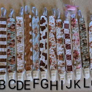 May include: A collection of fourteen fabric pacifier clips arranged in a row, each featuring a unique pattern including highland cows, floral prints, cow spots, and stripes. Each strap has a white plastic clip at the bottom and a loop at the top.