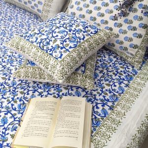 May include: Two square throw pillows with a white base, blue floral patterns, and green leaf borders, resting on a matching bedspread. An open book with printed text in a non-Latin script lies in the foreground, displaying pages of black text.