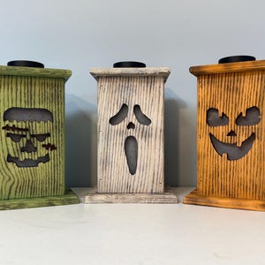 May include: Three wooden Halloween candle holders with different spooky faces carved into them. The candle holders are painted in green, white, and orange.