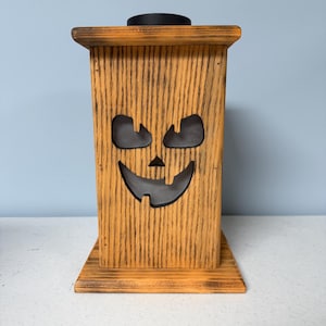 May include: A wooden jack-o'-lantern lantern with a black top and a cutout face. The lantern is stained in a light brown color and has a distressed finish.