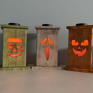 May include: Three wooden candle holders with different Halloween-themed faces. The faces are lit from behind with orange light. The candle holders are painted in green, gray, and brown.