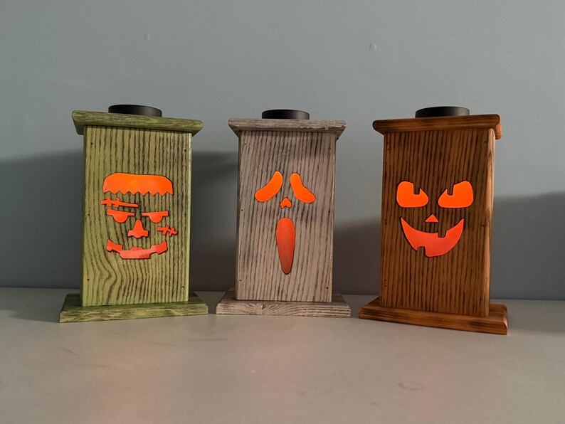 May include: Three wooden candle holders with different Halloween-themed faces. The faces are lit from behind with orange light. The candle holders are painted in green, gray, and brown.