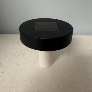 May include: A black solar powered light with a white base. The light has a square solar panel on top.