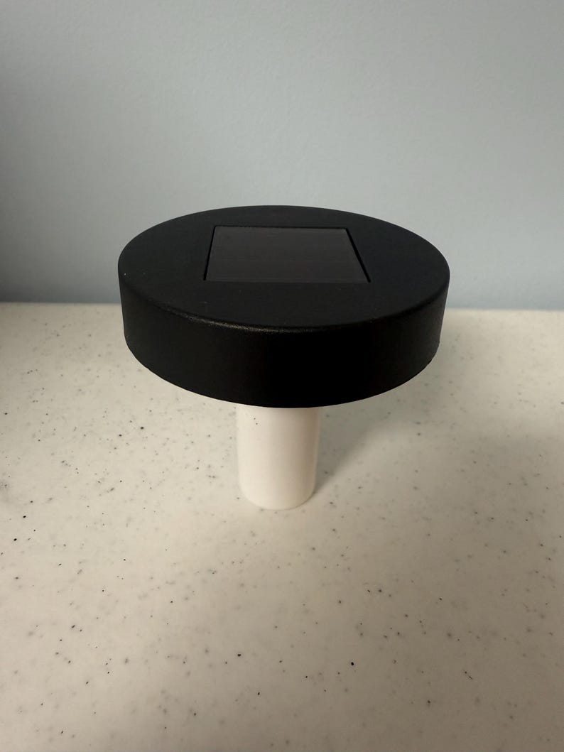 May include: A black solar powered light with a white base. The light has a square solar panel on top.