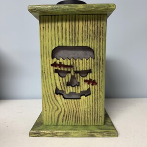 May include: Green wooden lantern with a cutout of a Frankenstein face. The lantern has a distressed finish and is perfect for Halloween decor.