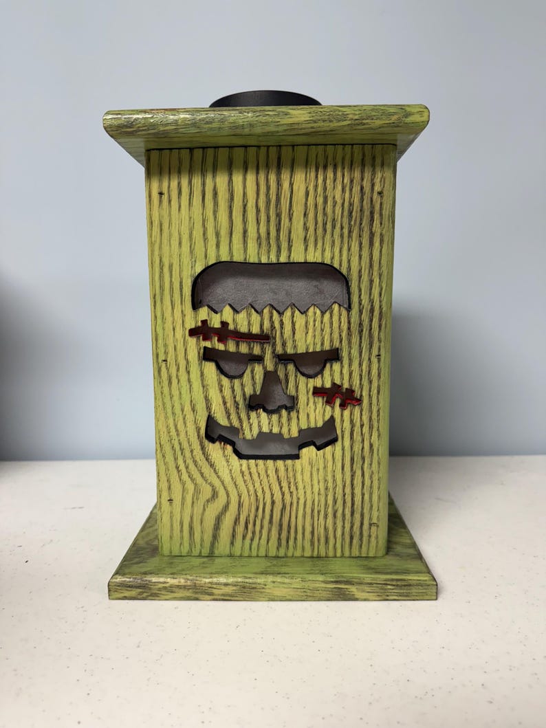 May include: Green wooden lantern with a cutout of a Frankenstein face. The lantern has a distressed finish and is perfect for Halloween decor.