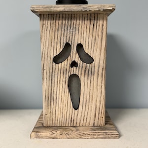 May include: A wooden candle holder with a ghost face cutout. The holder is painted in a light wood stain with a distressed finish.