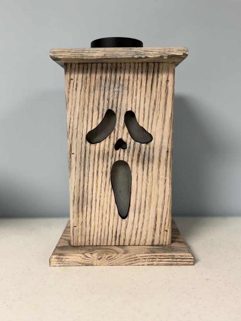 May include: A wooden candle holder with a ghost face cutout. The holder is painted in a light wood stain with a distressed finish.