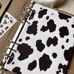 May include: A beige leather-textured ring binder featuring a translucent divider with a black and white cow print pattern. The binder includes silver metal rings and is accompanied by a clear pouch containing small circular stickers and a black tool.