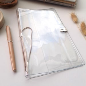 Puede incluir: Una funda transparente de plástico para cuaderno con un cierre de presión metálico y un bolígrafo a juego de color melocotón claro. La funda es rectangular con esquinas redondeadas y un acabado liso y transparente para proteger.