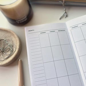 Könnte beinhalten: Ein weißer Papierplaner mit einem Rasterlayout, das Spalten für Montag, Dienstag, Mittwoch und Donnerstag enthält. Ein hellrosa Stift, eine kleine beige Schale mit silbernen Büroklammern und eine Glaskerze sind in der Nähe platziert.