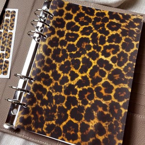 Può includere: Un raccoglitore beige effetto pelle con un meccanismo metallico a sei anelli e un inserto rimovibile con un motivo leopardato. Il raccoglitore include una tasca laterale contenente strisce adesive leopardate abbinate e una busta di plastica.