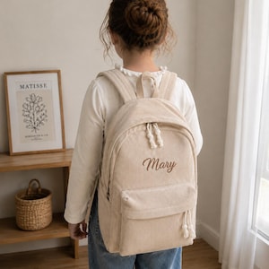 Peut inclure: Un sac à dos en velours côtelé beige avec une poche avant et le prénom Mary brodé en écriture cursive marron. Le sac possède deux bretelles, une poignée supérieure et des tirettes de fermeture éclair blanches à nœuds.