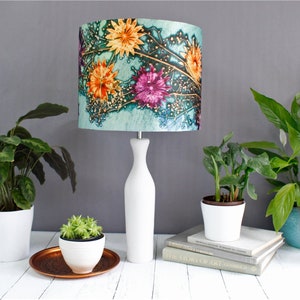 May include: A white table lamp with a floral patterned lampshade. The lampshade has a teal background with orange, pink, and purple flowers. The lamp is on a white table with two potted plants and a stack of books.