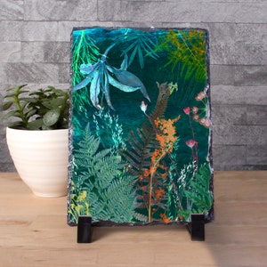 May include: A rectangular slate art piece with a botanical design. The artwork features ferns, flowers, and foliage in green, orange, and blue hues. The piece is supported by a black stand. The overall aesthetic is reminiscent of a vibrant, natural scene.