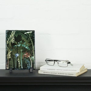 May include: A slate coaster with a pressed flower design featuring green, white, and red flowers. The coaster is on a black shelf with two books and a pair of glasses.