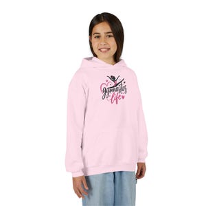 May include: A light pink hooded sweatshirt featuring a graphic on the chest. The design includes a black silhouette of a gymnast in a leap, a pink heart outline, and the text gymnastics life in a black script font with small pink stars.