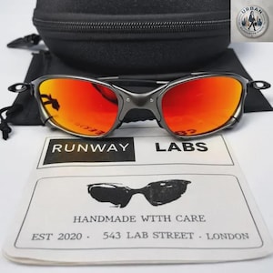 May include: Sunglasses with dark grey frames and vibrant orange-red reflective lenses. The set includes a black zippered hard case and a white microfiber cleaning cloth printed with the text RUNWAY LABS, a graphic of sunglasses, and address details.