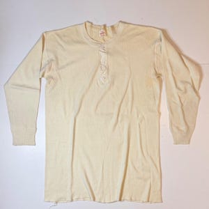 May include: A long-sleeve henley shirt in a solid off-white colour. The garment features a round neckline with a vertical placket containing three matching buttons. The sleeves have ribbed cuffs, and a small white label is visible at the inner neck.
