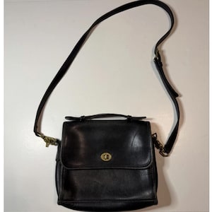 May include: A black leather crossbody bag featuring a top handle, a fold-over flap with a brass-toned turn-lock closure, and an adjustable shoulder strap attached with brass-toned metal hardware clips on both sides of the structured bag.