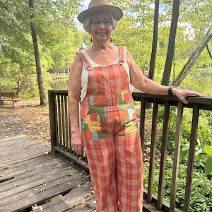 May include: A pair of orange plaid overalls featuring patchwork pockets with multi-colored floral and geometric patterns, worn over a white sleeveless knit top, paired with a straw hat, glasses, and black thong sandals.
