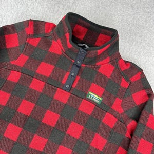 May include: A red and black buffalo plaid fleece pullover featuring a mock neck collar with a dark navy blue snap-button placket. The garment includes a small rectangular L.L.Bean brand patch on the chest with green, blue, and yellow accents.