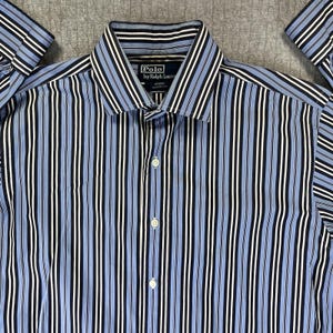 Op de afbeelding: Een overhemd met lange mouwen en knoopsluiting met een verticaal gestreept patroon in lichtblauw, marineblauw, zwart en wit. Het overhemd heeft een puntige kraag en een marineblauw binnenlabel met de tekst Polo by Ralph Lauren, Regent Classic Fit.
