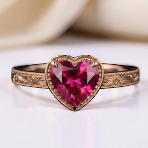 Könnte beinhalten: Ein Ring aus Roségold mit einem herzförmigen, tiefrosa Edelstein in der Mitte. Die Ringschiene ist an den Seiten mit filigranen, gravierten Rankenmustern verziert, und der Stein ist von einem zarten, perlenartigen Metallrand umgeben.