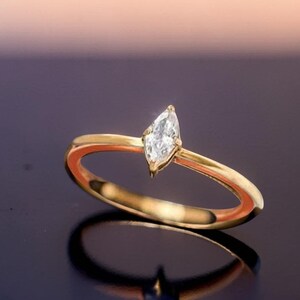 Könnte beinhalten: Ein goldener Ring mit einem einzelnen, klar geschliffenen Edelstein im Marquise-Schliff, der in eine schlichte, polierte Ringschiene gefasst ist. Der Ring ist so positioniert, dass die schlanke, längliche Form des Steins sichtbar wird.