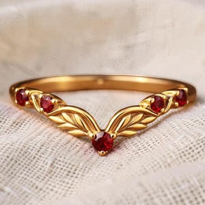 Könnte beinhalten: Ein goldener V-förmiger Ring mit einem zentralen runden roten Edelstein, flankiert von zwei geformten Blattmotiven auf jeder Seite, sowie zwei weiteren runden roten Edelsteinen an den äußeren Enden, was ein symmetrisches Blattdesign ergibt.